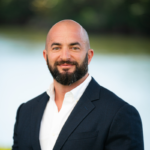 Confident man in a dark suit smiling outdoors by water.