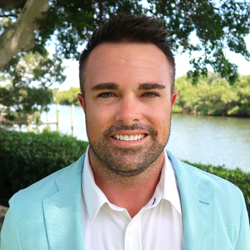 A smiling man in a light blue blazer outdoors by a river.