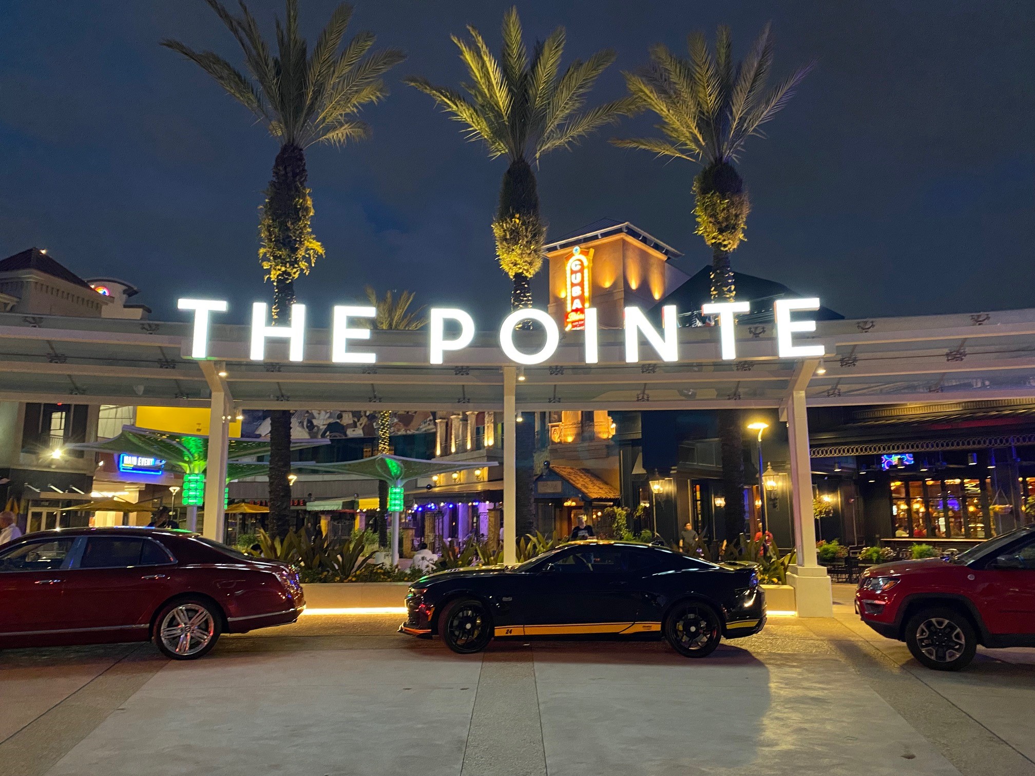 Night view of The Pointe with parked cars and illuminated palm trees.
