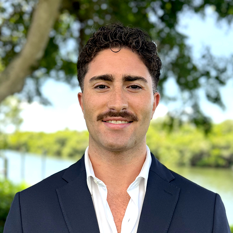 Smiling man in a suit standing outdoors with trees and water in the background.