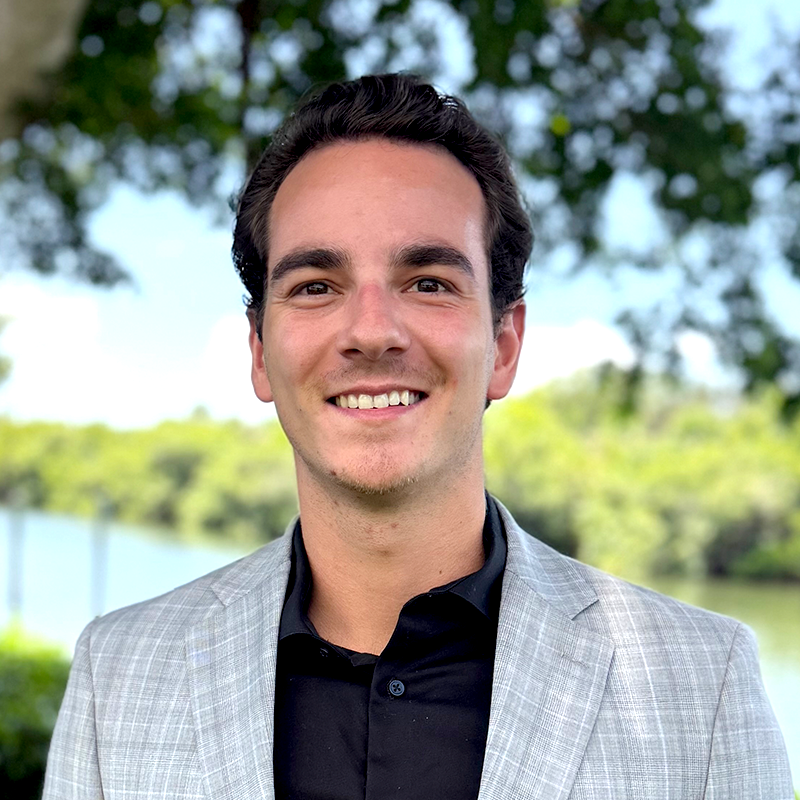 A smiling man in a light gray blazer with a black shirt outdoors.